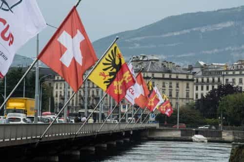 Verschiedene Schweizer Flaggen auf einer Brücke aneinandergereiht; daneben Verkehr und Spaziergänger.