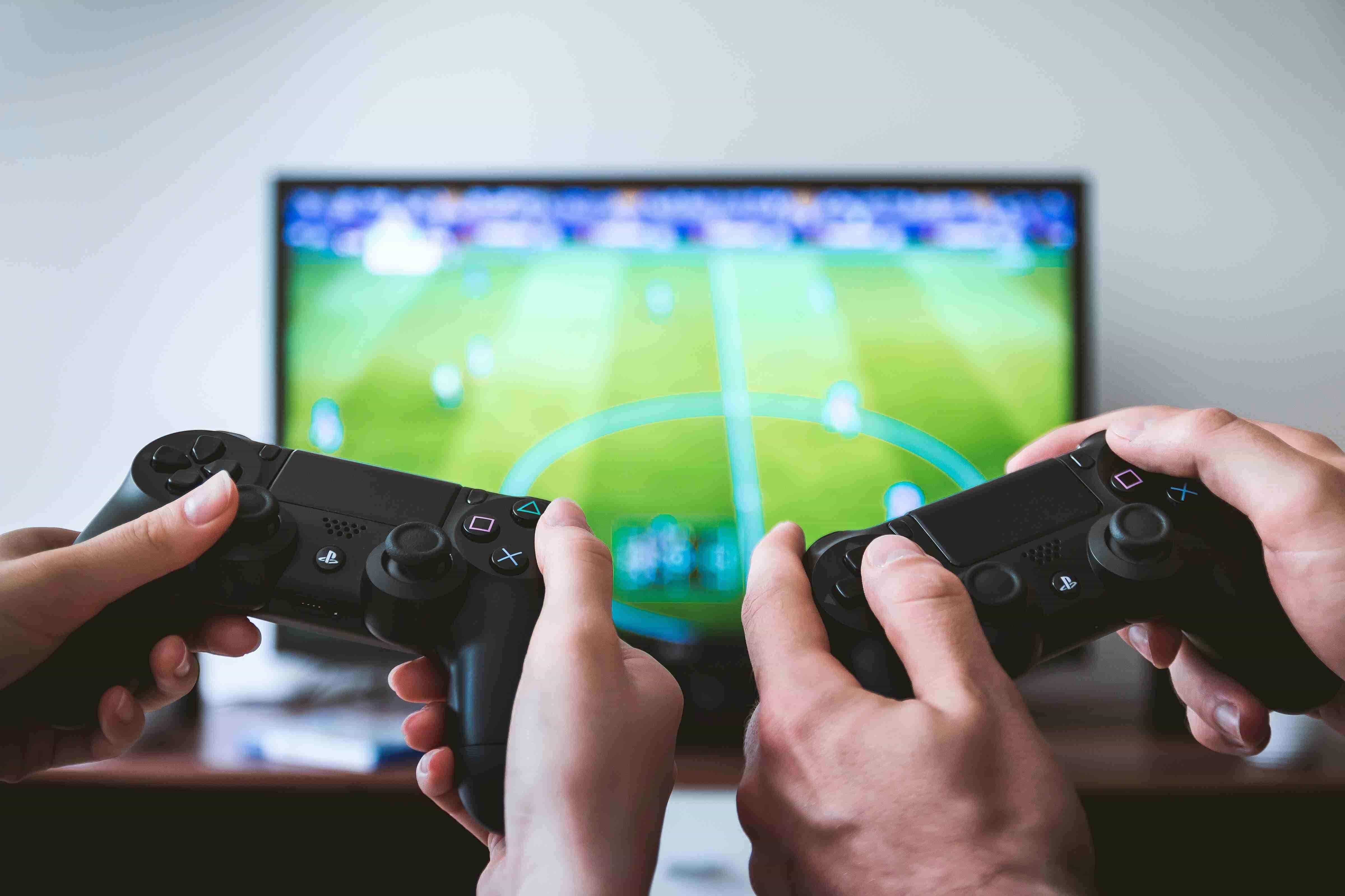 Dua pengontrol Playstation di tangan seorang pria dan wanita yang tampak sedang bermain FIFA, buram di TV di latar belakang.