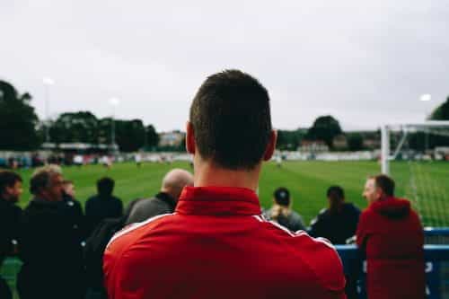 Ein Mann in rotem Sportanzug dreht den Rücken zur Kamera, steht im Stadion auf der Tribüne und beobachtet ein Fußballspiel zusammen mit anderen Fans.