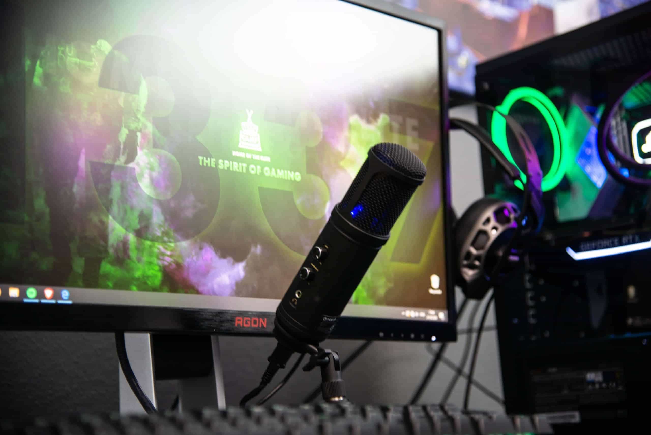 Auf einem Schreibtisch ist unterschiedliches aktiviertes Streaming-Equipment zu sehen, wie beispielsweise ein Standmikrofon, Kopfhörer und ein Monitor
