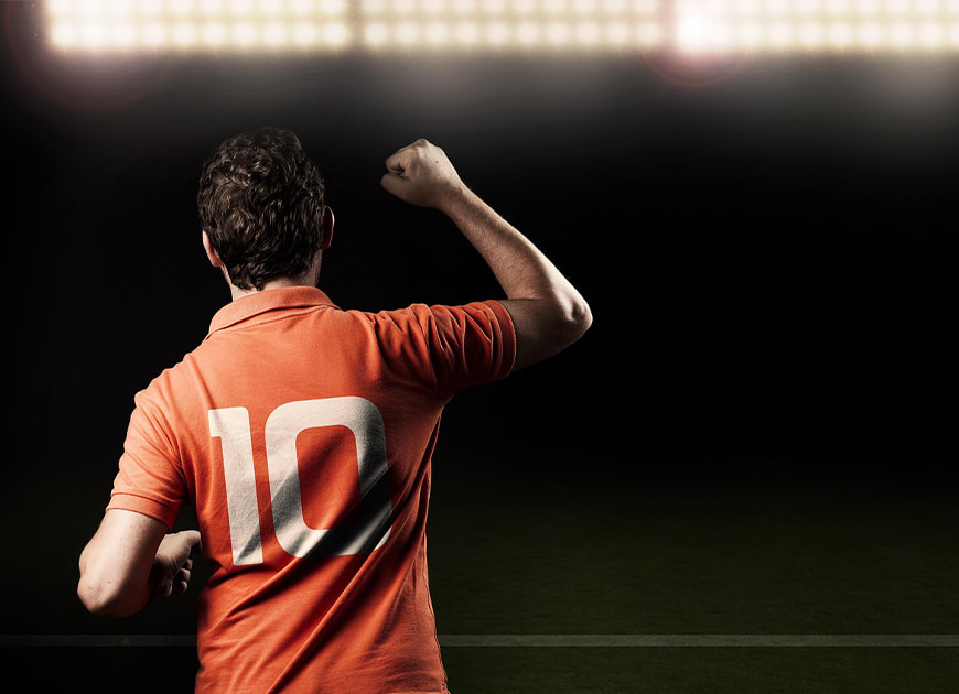 Ein jubelnder Fußballer im orangefarbenen Trikot mit der Nummer 10 steht unter grellem Stadionlicht vor dunklem Hintergrund.